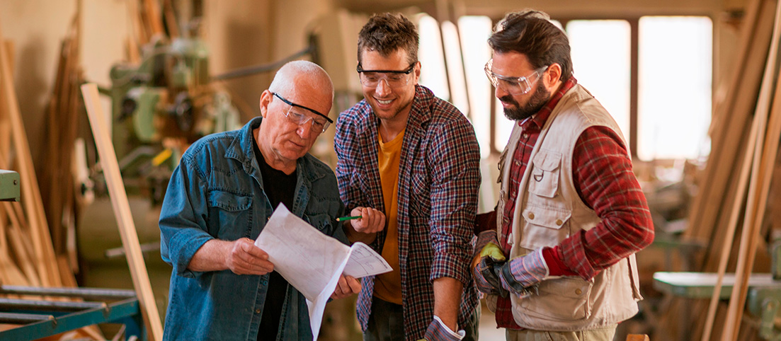 Traja muži v dielni s ochrannými okuliarmi diskutujú nad plánmi na papieri.