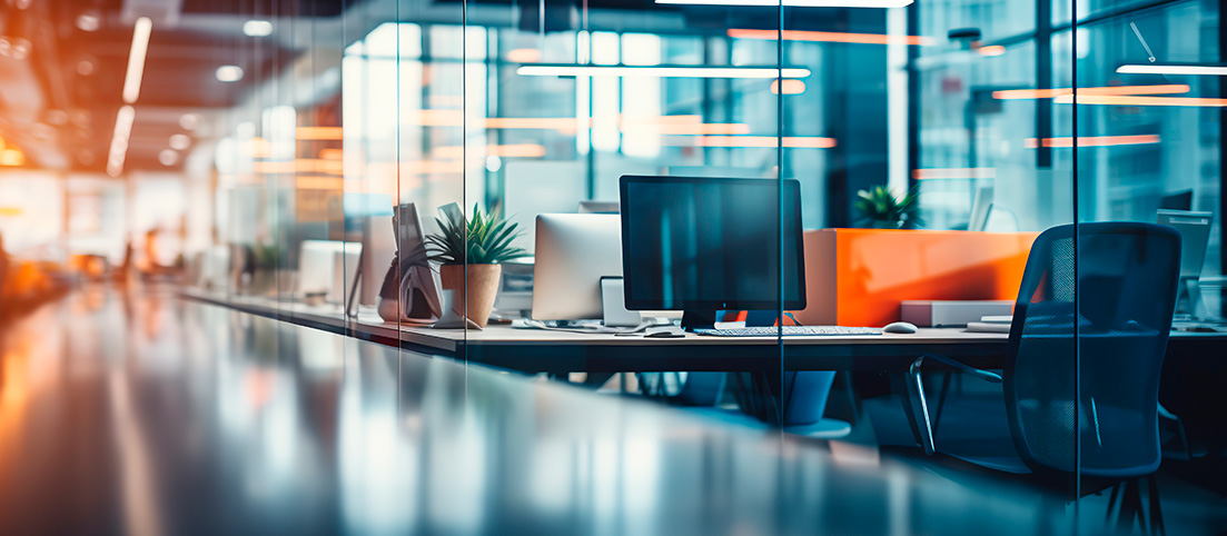 Moderná kancelária s počítačmi, kancelárskymi stoličkami a rastlinami za sklenenými priečkami.
