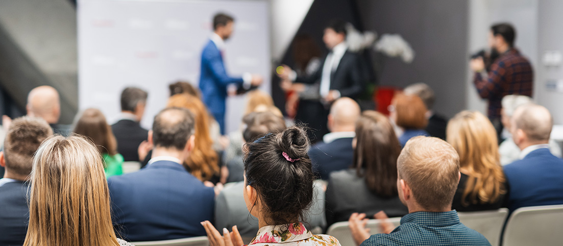 Ľudia sediaci v sále sledujú odovzdávanie ceny medzi dvoma mužmi v pozadí.