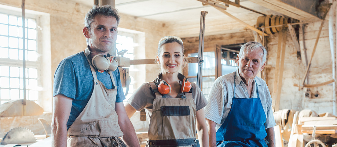 Trojica rodinných členov v dielni, muž, žena a starší muž v pracovnom oblečení s ochrannými slúchadlami.