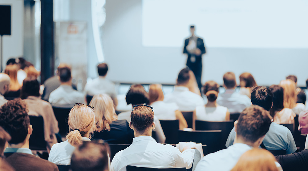 Ľudia sediaci v sále počúvajúci prednášajúceho počas biznis podujatia alebo konferencie.