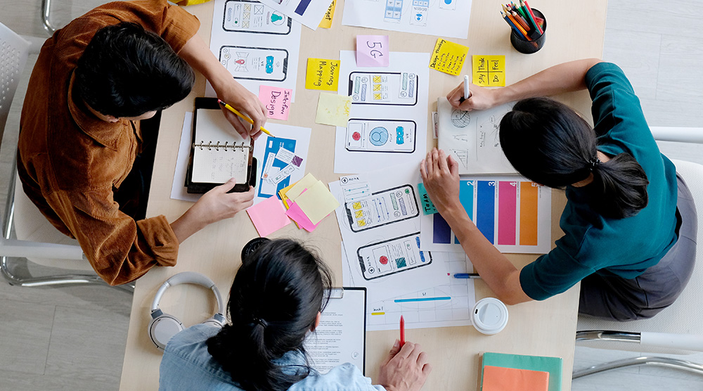 Tím ľudí pracuje na návrhu mobilnej aplikácie s náčrtmi, poznámkami a farebnými štítkami na stole.