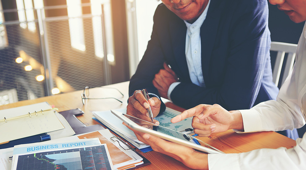 Dve osoby v oblečení diskutujú nad tabletom a obchodnou správou s grafmi na stole v kancelárii.