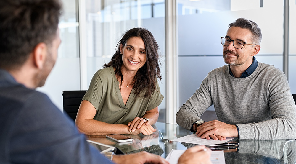 Dvojica klientov počúva odborné poradenstvo od konzultanta v kancelárii pri stole so zmluvami.