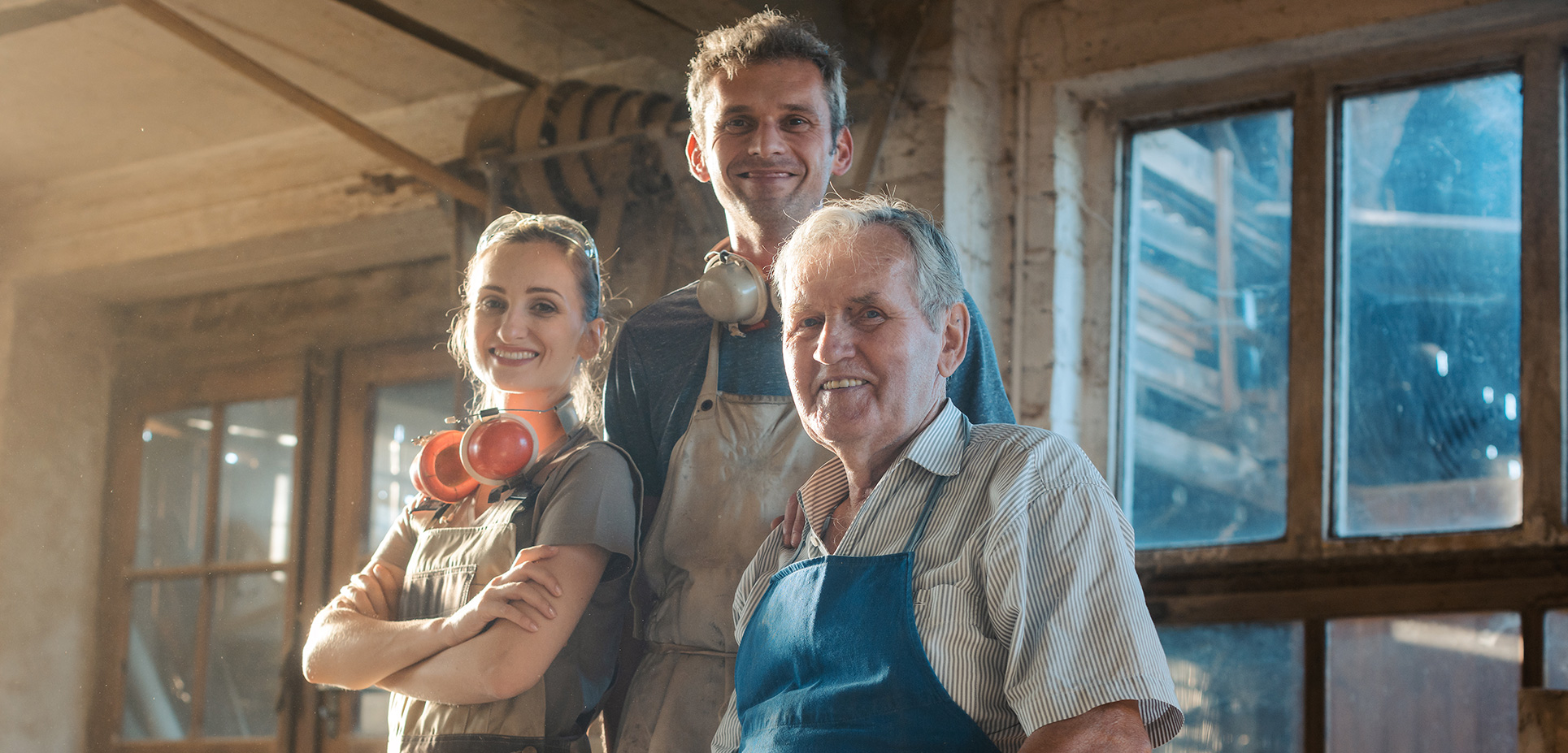 Trojica usmievajúcich sa ľudí v dielni, pravdepodobne rodinní remeselníci v pracovných odevách.