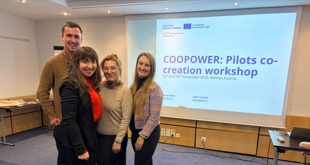 Štyria usmiati účastníci workshopu COOPower v zasadačke s premietacou plátnou v pozadí.