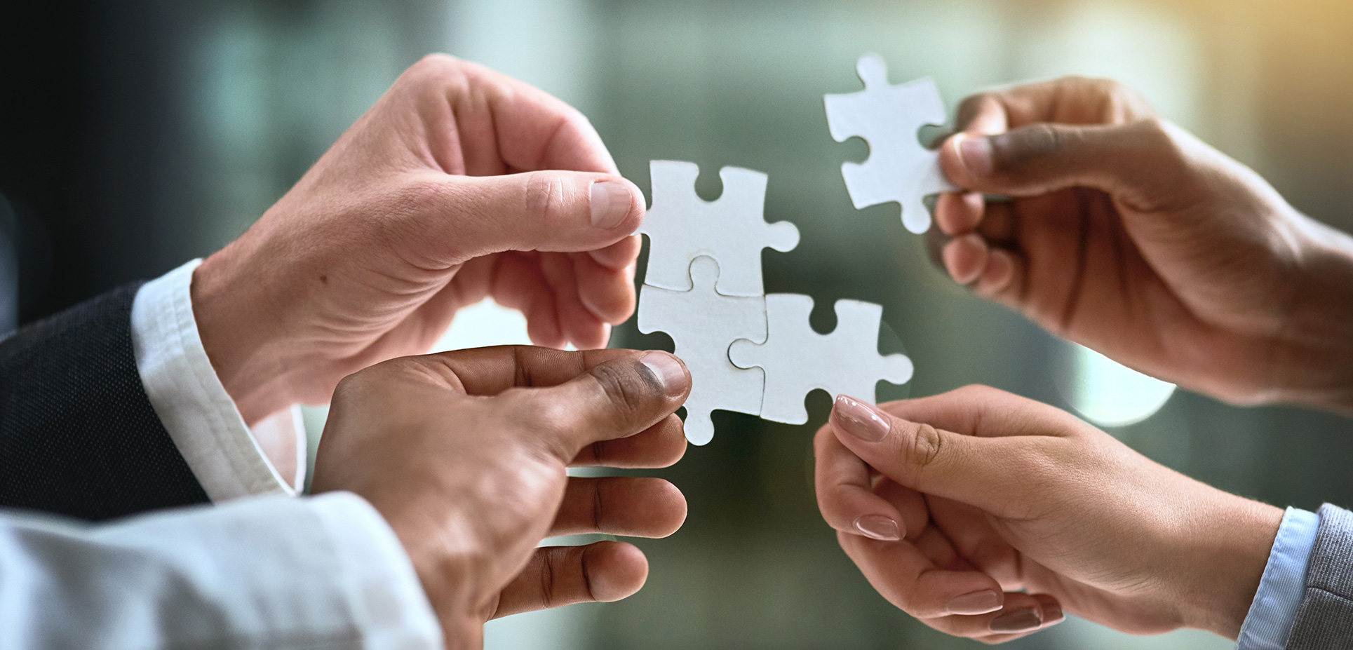 Štyri ruky rôznych ľudí skladajú biele puzzle symbolizujúce spoluprácu a riešenie problémov.