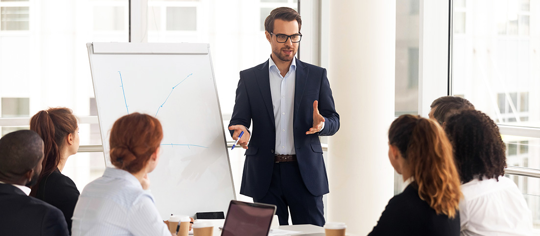 Muž v obleku vedie prezentáciu pred skupinou ľudí, na tabuli sú načrtnuté grafy.