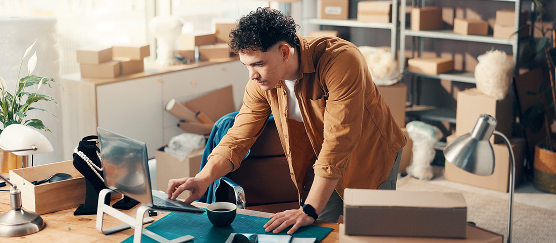 Mladý muž pracuje na notebooku v kancelárii plnej kartónových krabíc a balíkov, pravdepodobne malý podnikateľ.