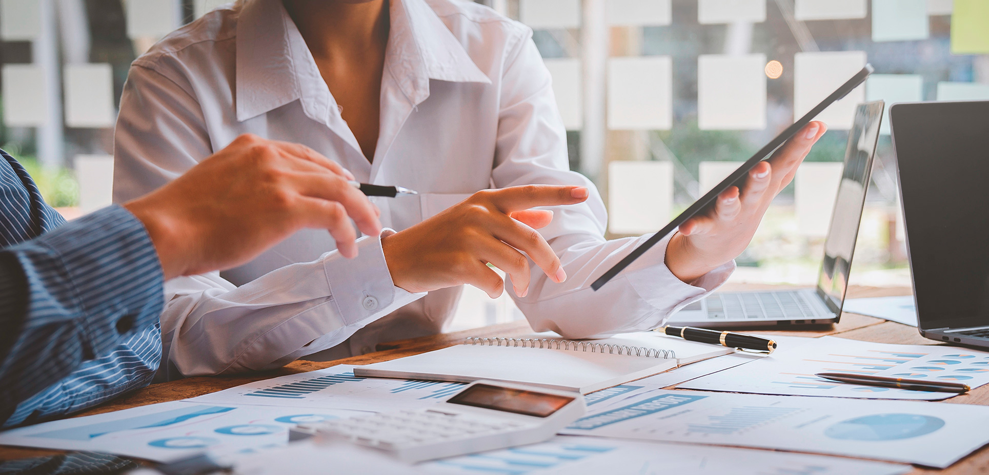 Dve osoby diskutujú nad tabletom a dokumentmi s grafmi a kalkulačkou na stole v kancelárii.