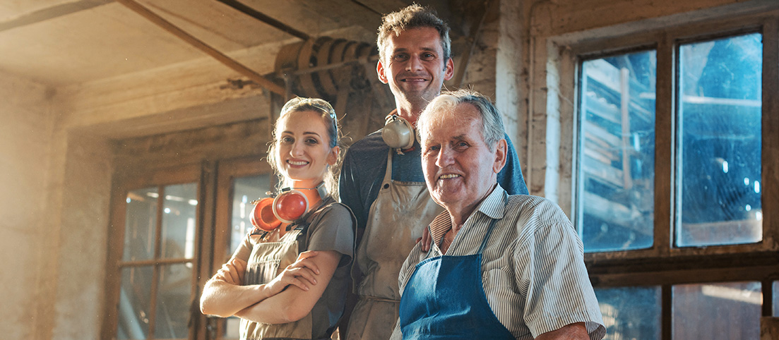 Trojica usmievajúcich sa remeselníkov v dielni, dvaja muži a žena v pracovnom odeve a ochranných slúchadlách.