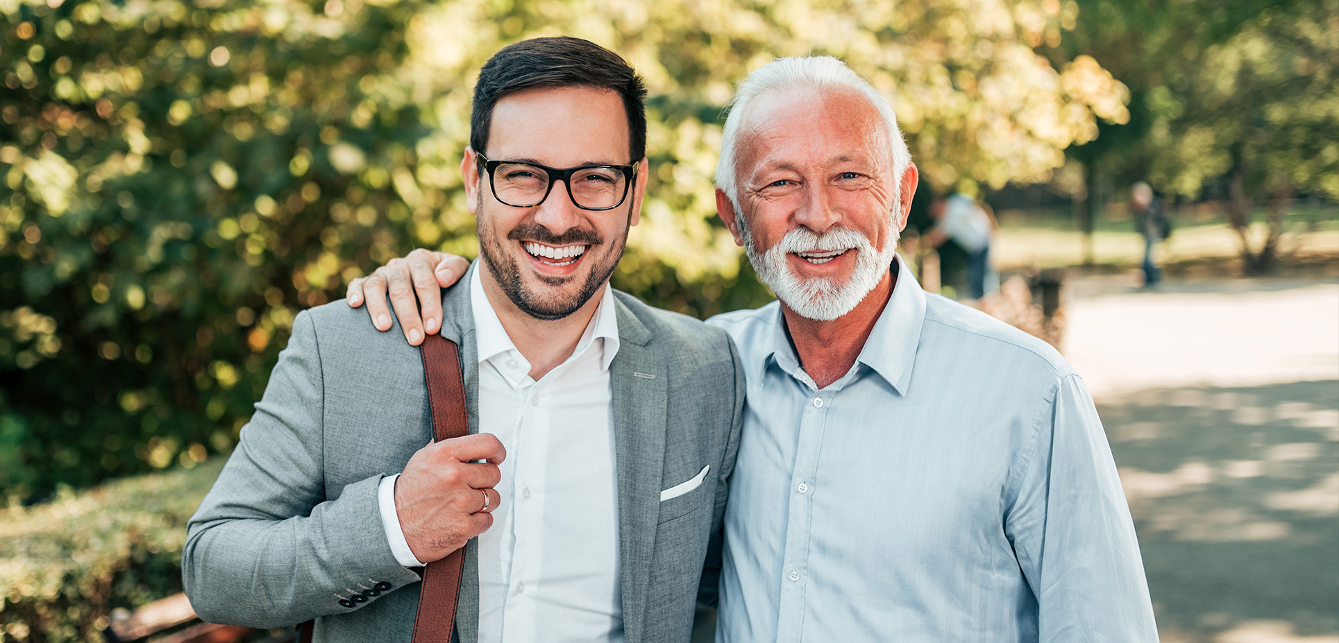 dve usmievajúce sa generácie mužov, mladý v obleku s taškou a starší s bielymi vlasmi v parku